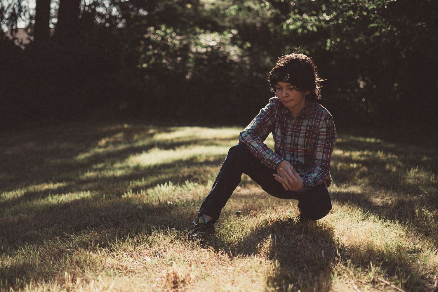 Teenager with curly hair crouching in dappled sunlight on grass, looking contemplative
