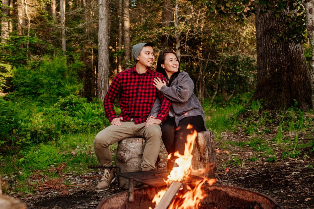 Two people sit close together by a warm campfire in a forest clearing during golden hour.