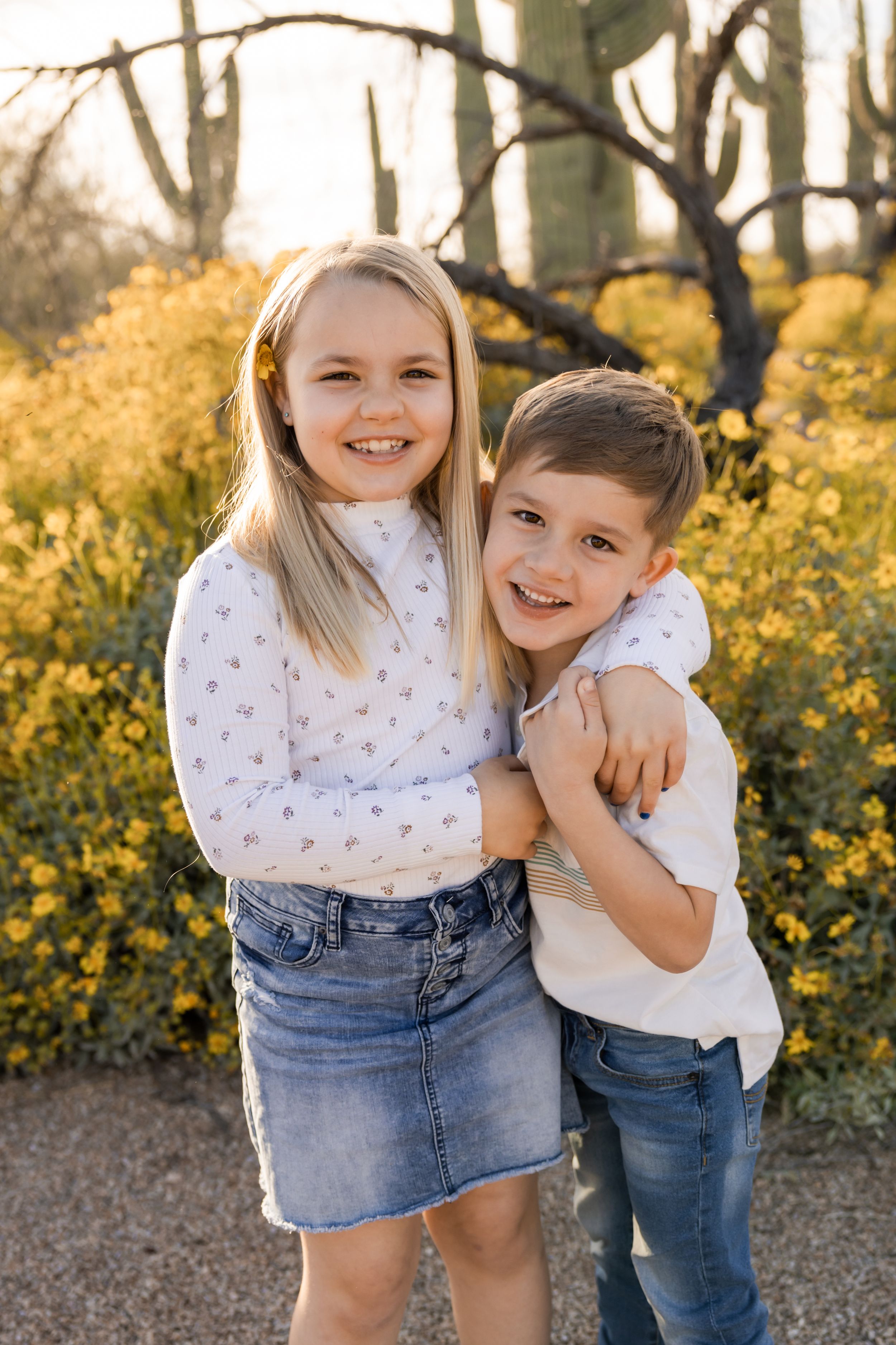 Two children hugging and smiling among yellow wildflowers at sunset.