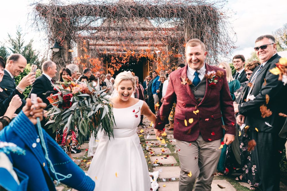 Joyful newlyweds walk through confetti shower hand in hand after outdoor autumn wedding ceremony.