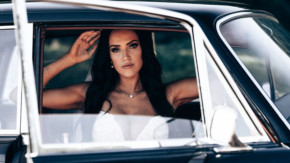 Elegant dark-haired woman in a white dress sits inside a vintage black car looking through the window.