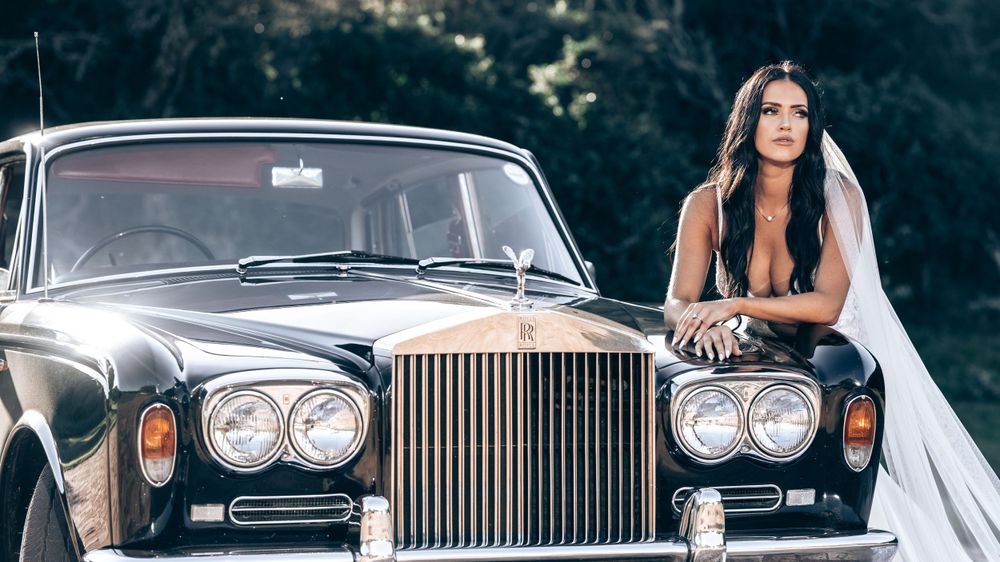 Bride in a wedding gown and veil poses leaning against a classic black Rolls-Royce outdoors.