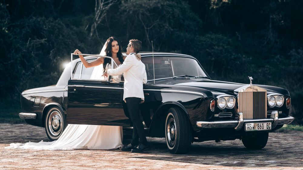 Bride and groom dance together beside a classic black Rolls-Royce in a lush outdoor setting.