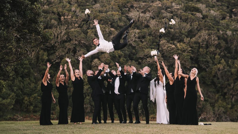 Wedding party in black formal wear tosses a groom into the air outdoors with hills behind them.