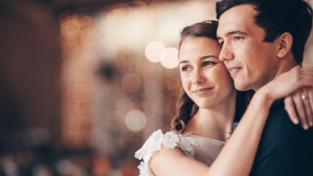 Romantic couple in wedding attire embrace and smile together in a warmly lit reception venue.