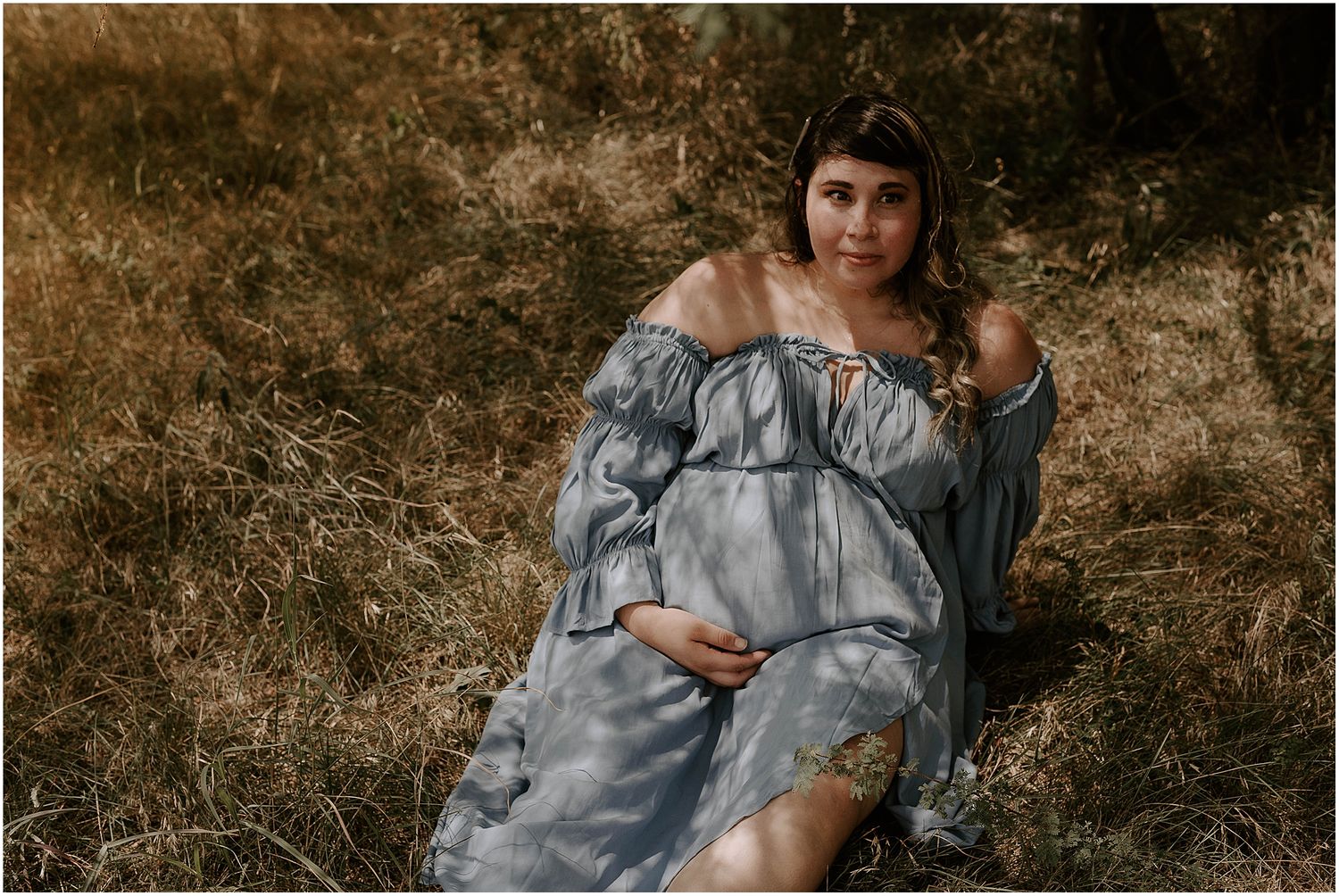 Maternity photo of an expectant mother in a flowing grey off-shoulder dress reclining in a natural outdoor setting.