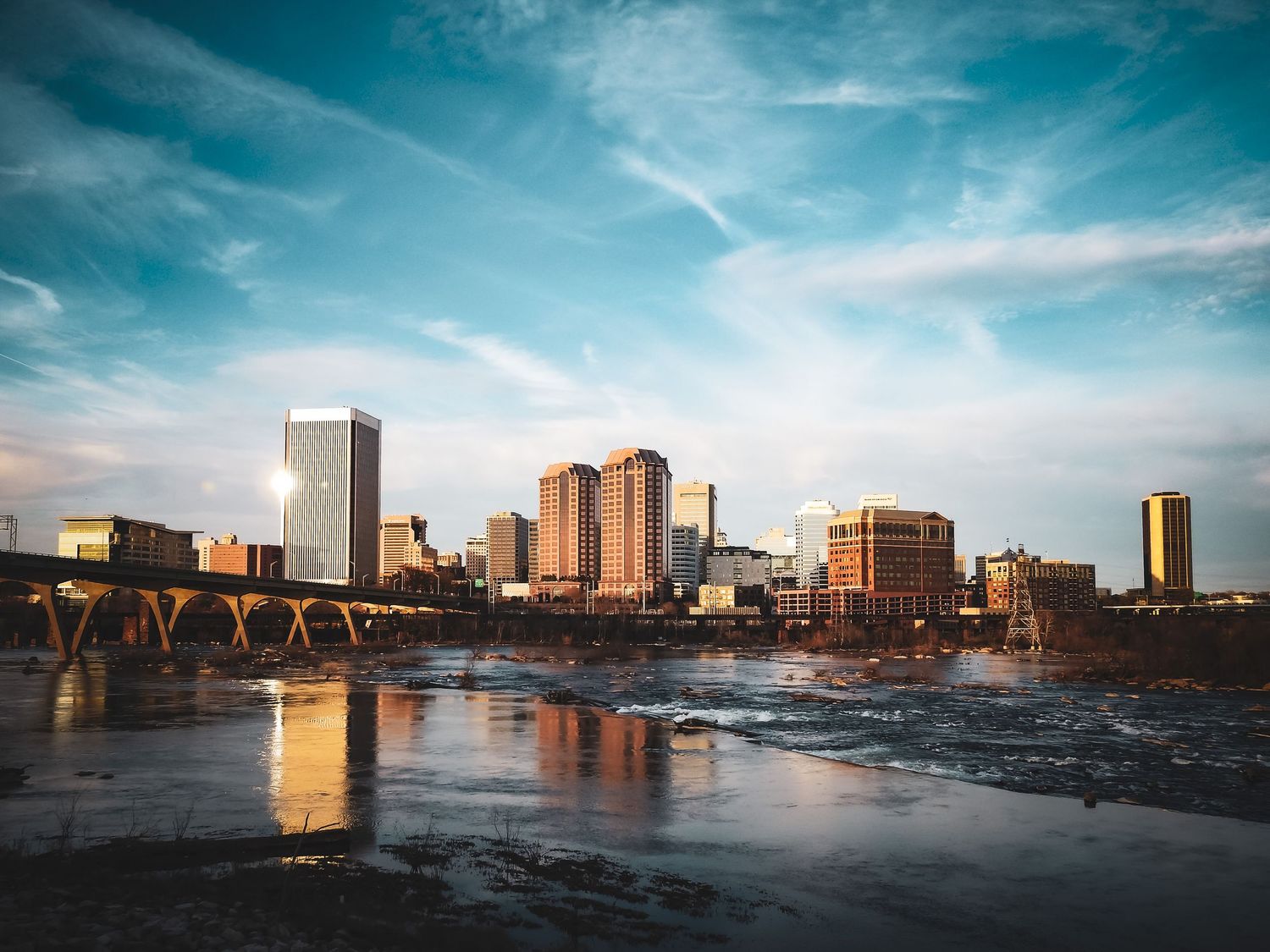 Richmond, Virginia skyline reflects in the James River at sunset with bridge and urban architecture.