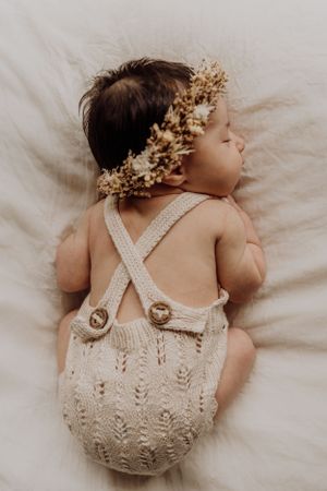 A sleeping baby wearing a knitted romper and floral crown photographed from above.