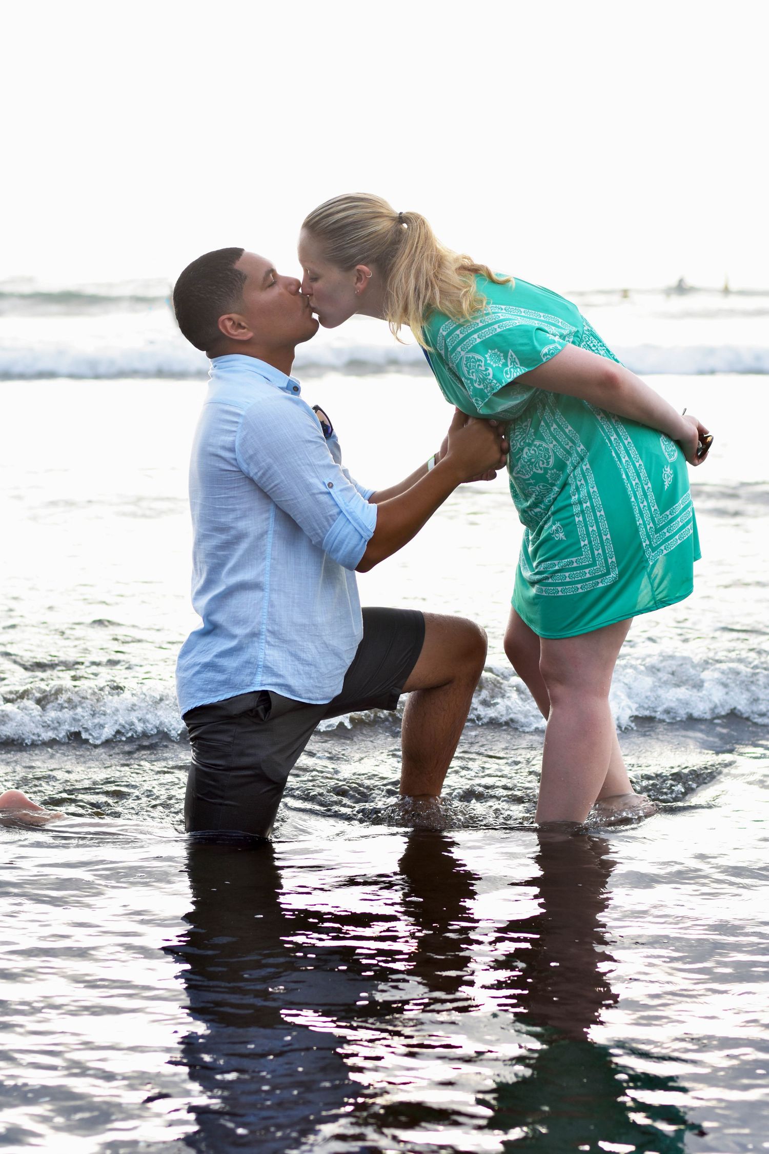 Man kneeling in the ocean kissing his fiancé