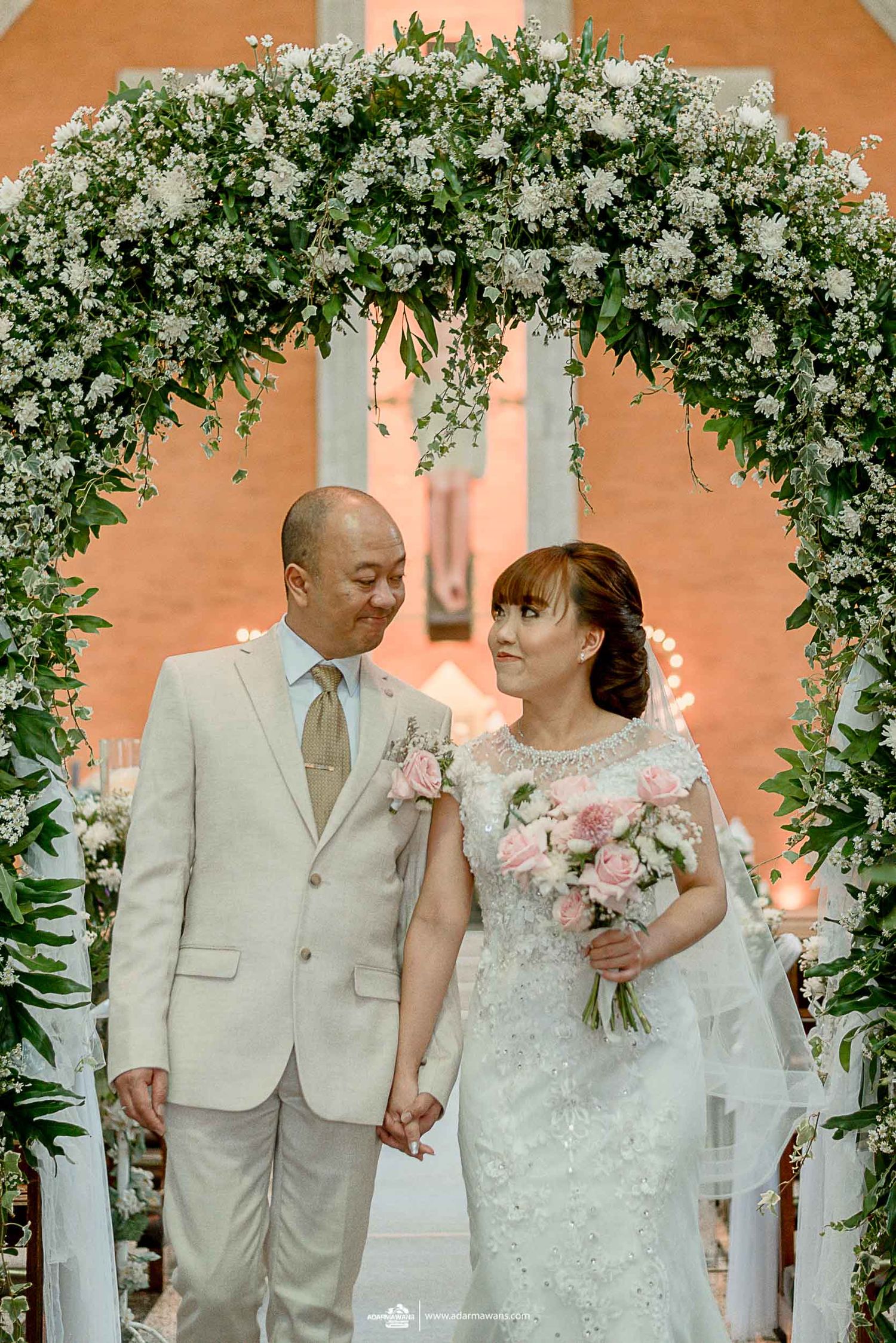 Wedding couple standing under floral arch with white flowers and greenery.