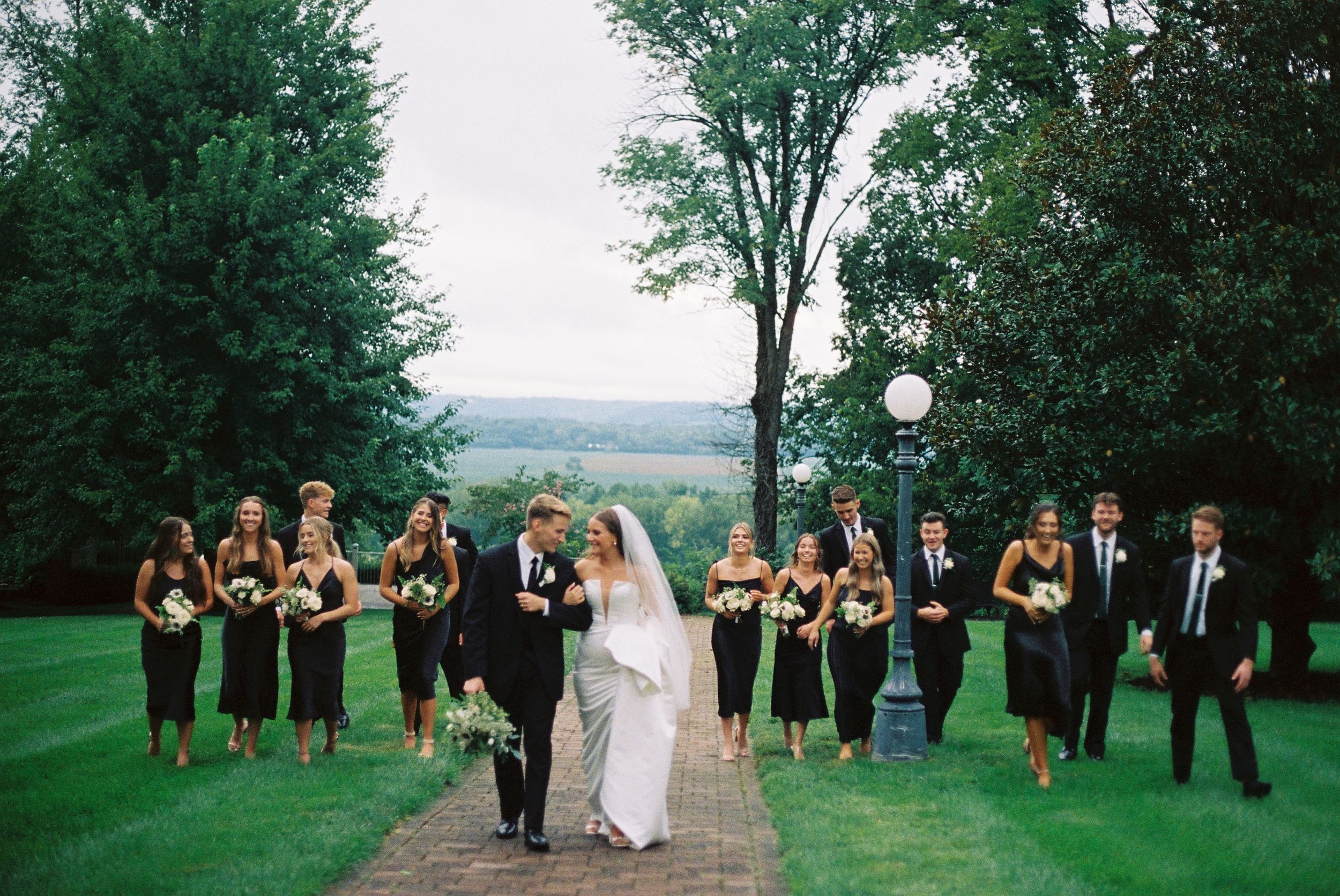 Wedding party lines up on a scenic outdoor path surrounded by green trees.