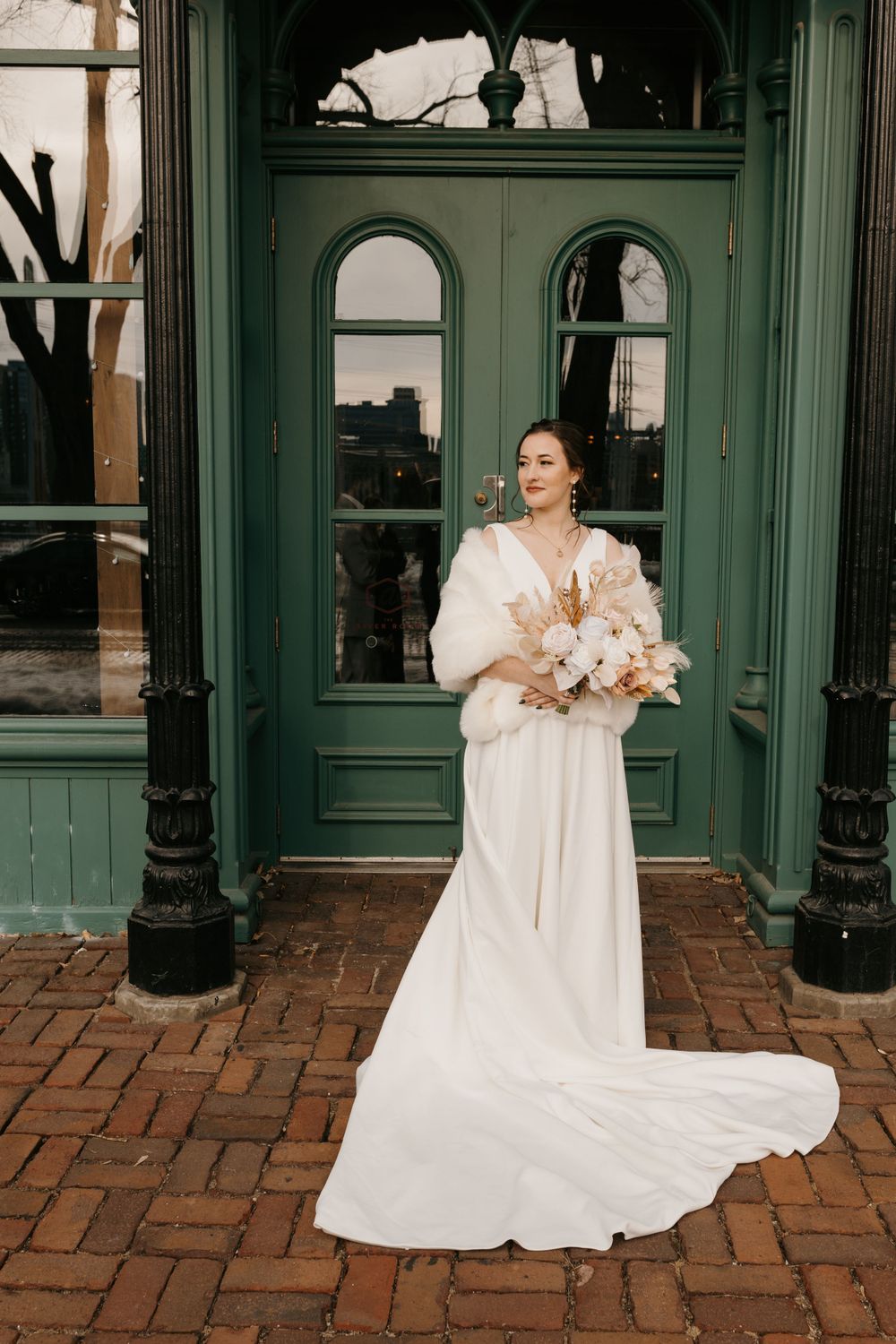 Bride in elegant white gown with fur stole holds dried floral bouquet before green vintage doors.