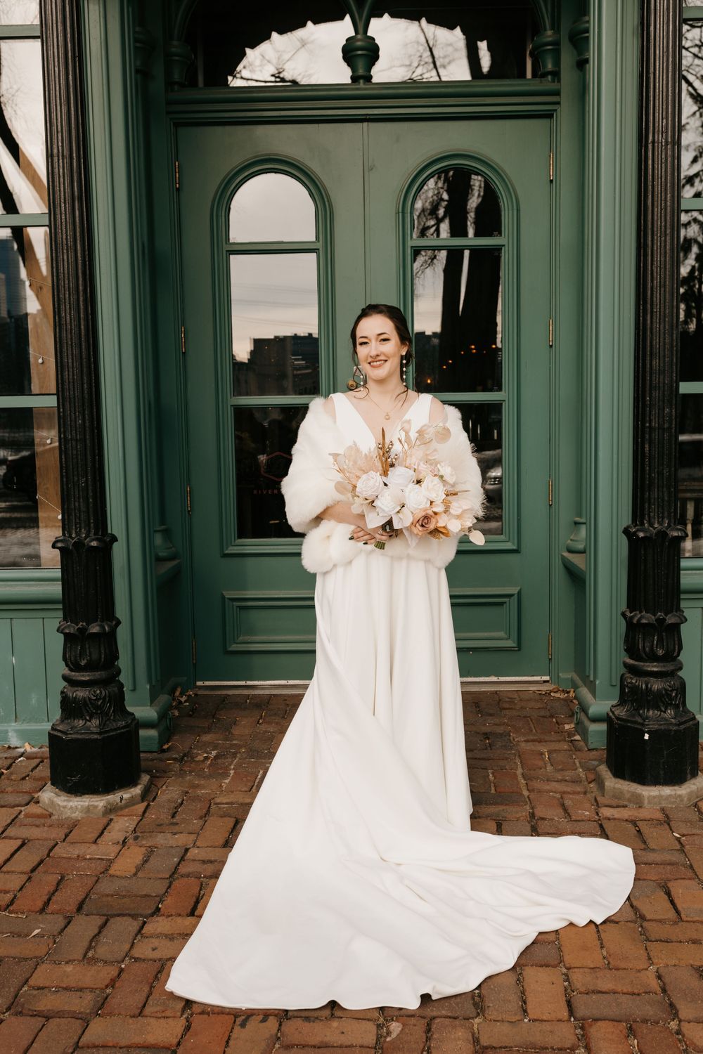 Smiling bride in white gown and fur wrap holds boho bouquet before green arched doors.
