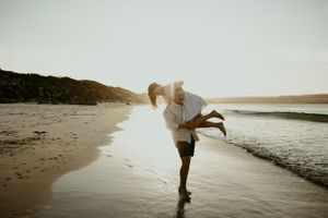A couple dances playfully on a scenic beach at sunset with flowing fabric creating a romantic silhouette.