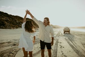 A couple shares a joyful dance moment on a coastal beach road at golden hour.
