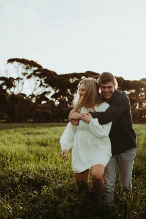 Couple embracing in a grassy field at sunset with trees in the background.