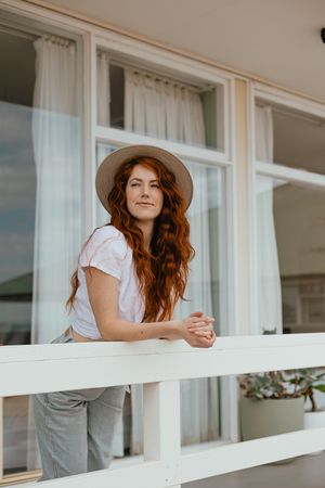 A person in a hat leans on a white porch railing with windows in the background.