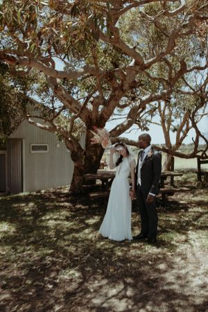 A wedding couple poses under a large shady tree during an outdoor ceremony.