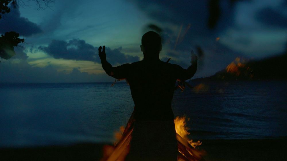 Silhouetted figure stands with raised arms near a beach bonfire at dusk with ocean in background.
