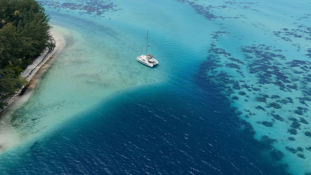 Aerial view of catamaran anchored between turquoise waters and sandy beach.