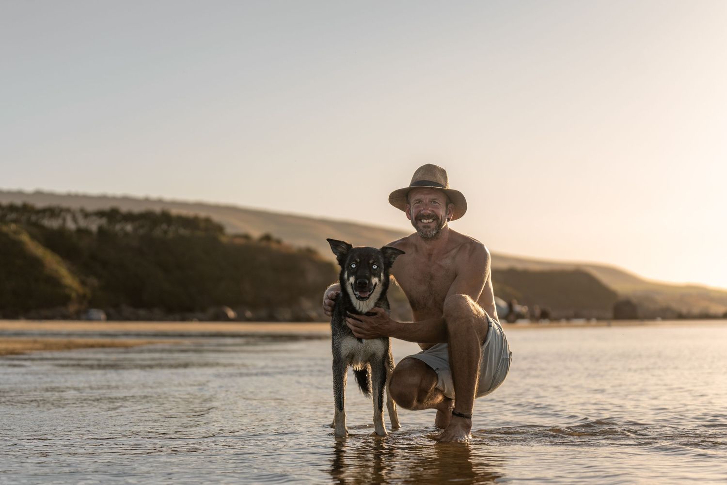 Smiling man in a hat crouching beside a black dog on a sunny beach at golden hour.