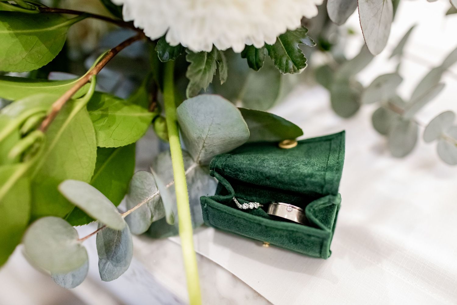 Close-up of wedding rings in a dark green velvet box nestled among white flowers and greenery.