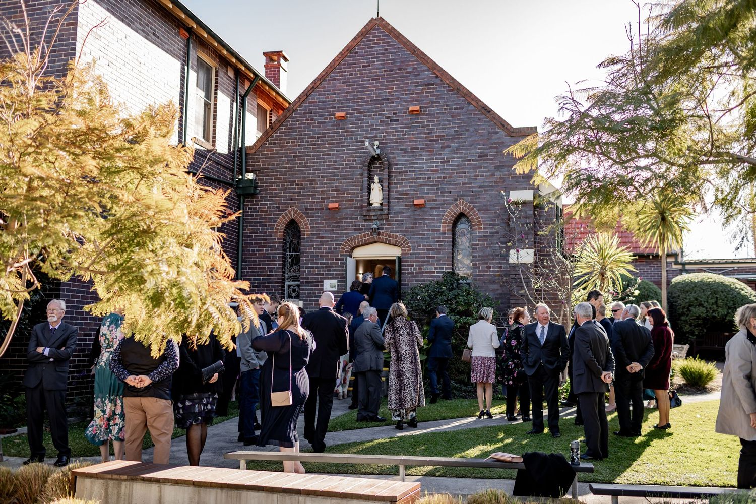 Wedding guests gather outside a stone church building on a sunny day.
