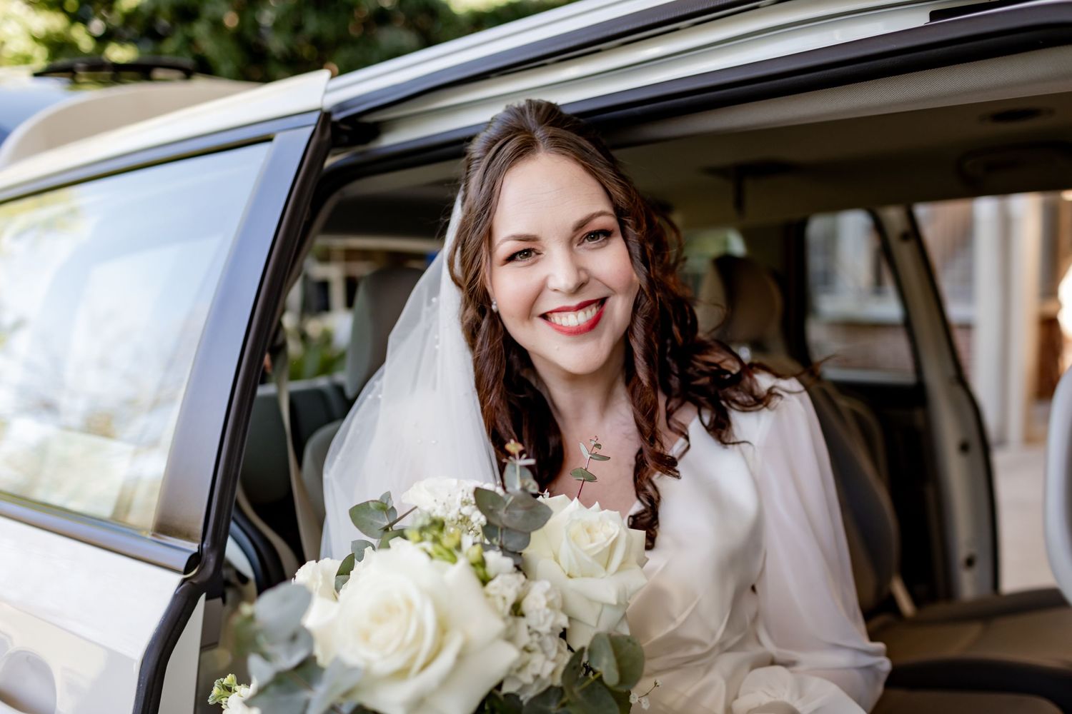 A bride in white dress sits in car holding bouquet of white flowers.