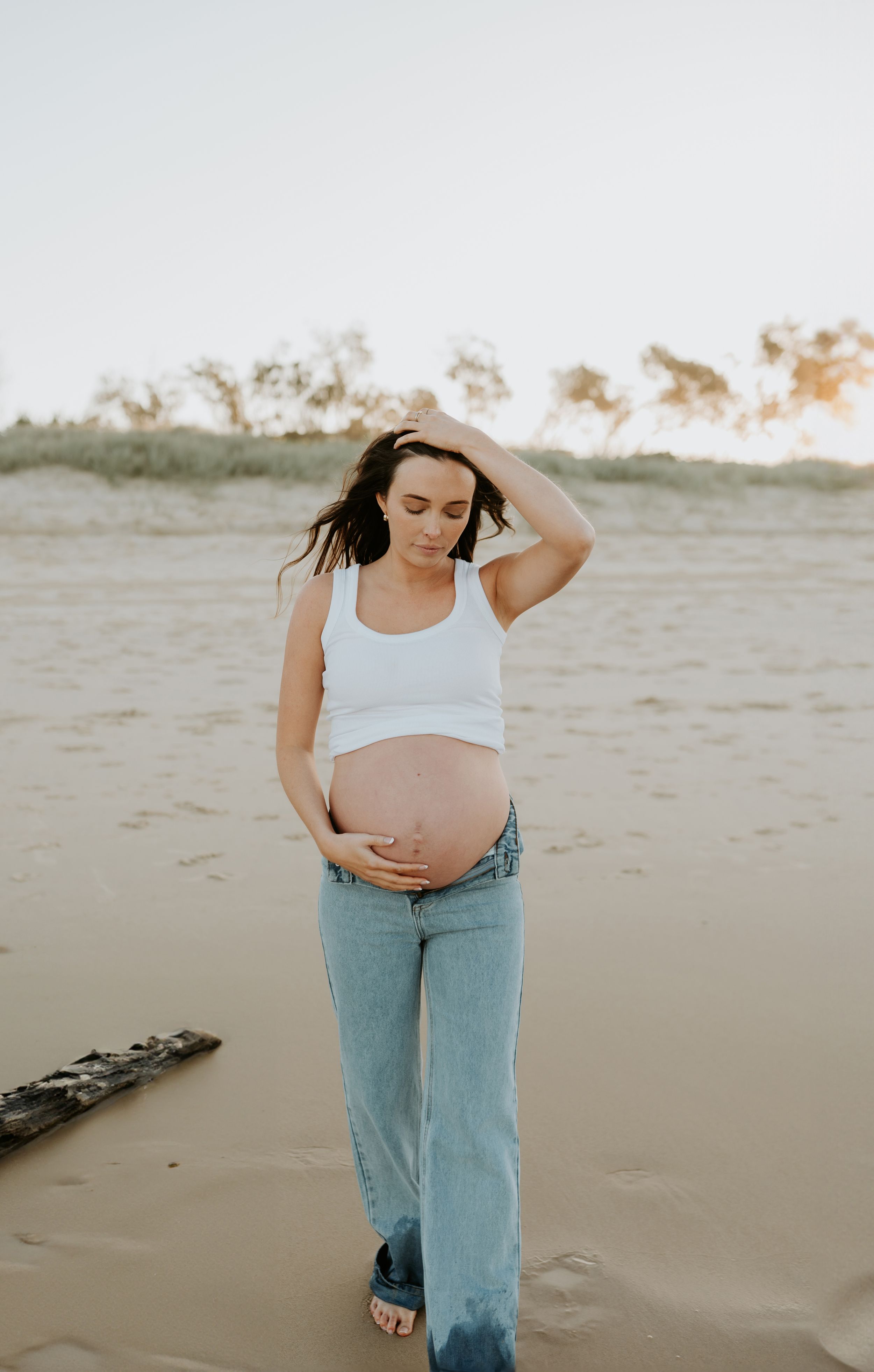 Expectant mother in casual attire stands thoughtfully on sandy beach during sunset.