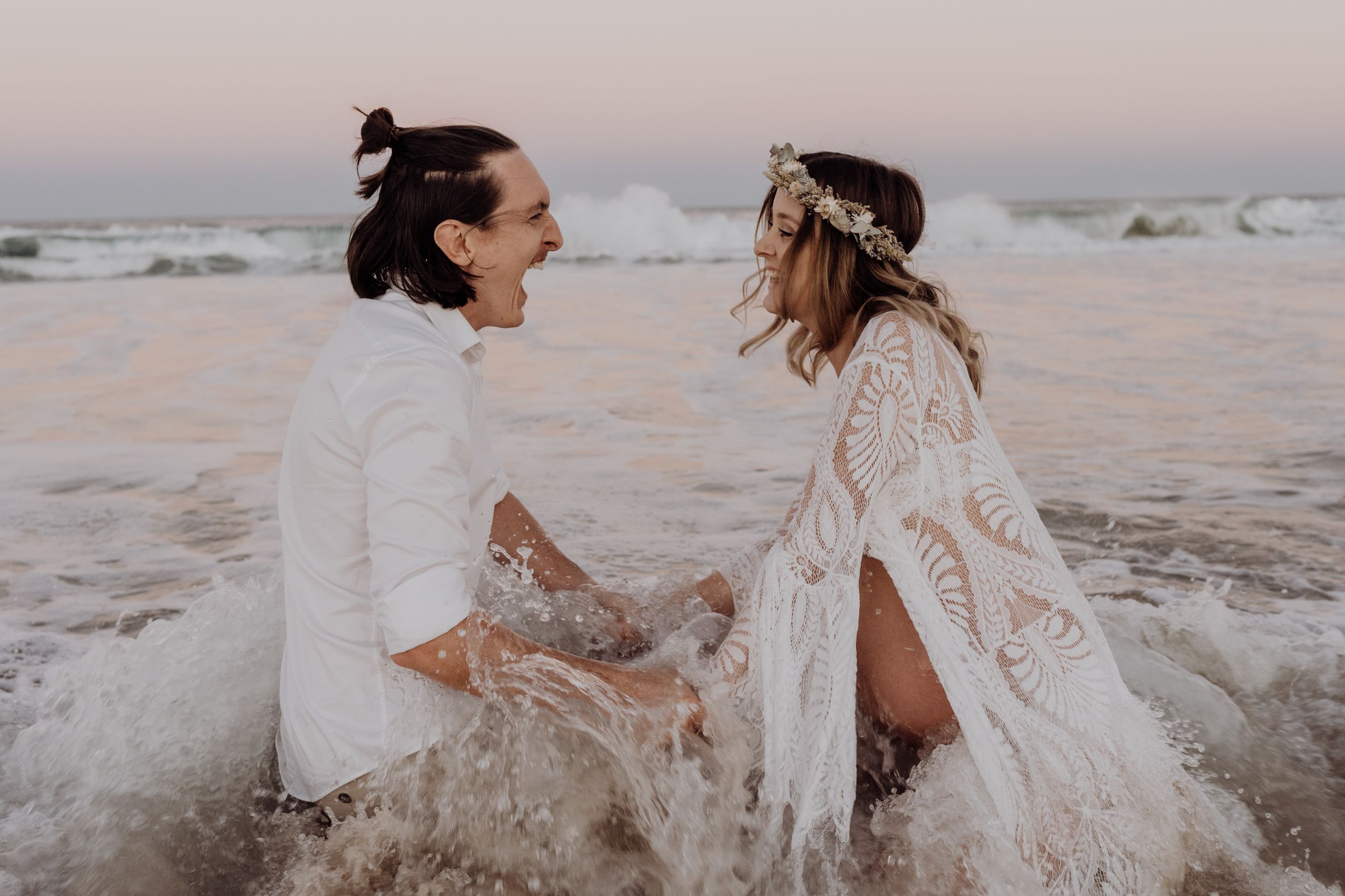 Two people in white outfits dancing and playing on the beach at dusk.