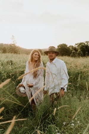 A couple poses together in tall grass during golden hour wearing light colored clothing.