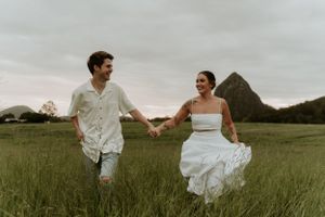 A joyful couple runs hand in hand through a green field with mountains in background.