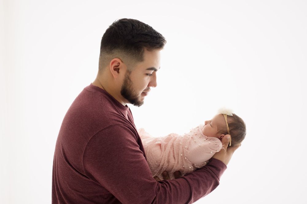 Parent gazing lovingly at newborn in pink outfit against white backdrop.