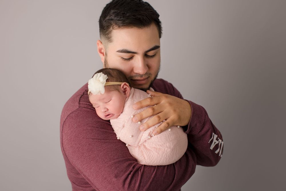 Parent in burgundy sweater gently cradling sleeping newborn wrapped in soft pink.