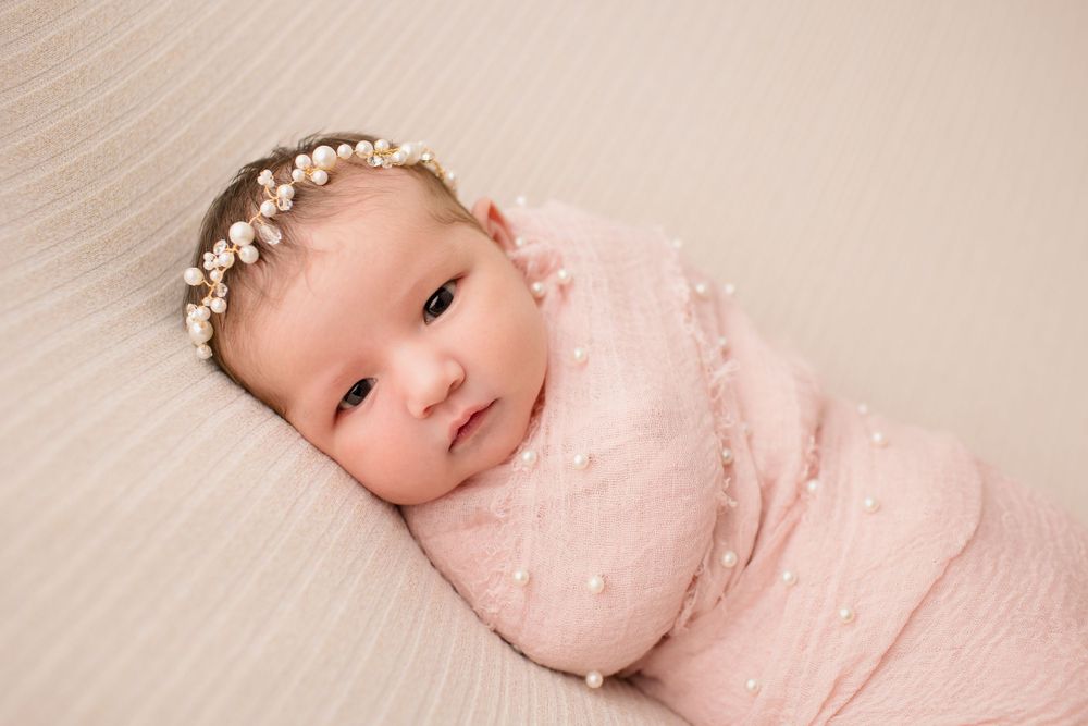 Newborn baby wearing pearl headband and pink swaddle on textured backdrop.