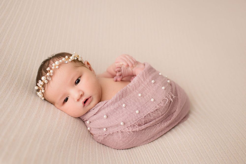 Sweet newborn portrait with pearl crown and pink wrap on cream textured background.