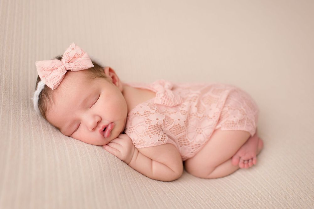 Sleeping newborn in pink lace outfit with bow headband on cream backdrop.