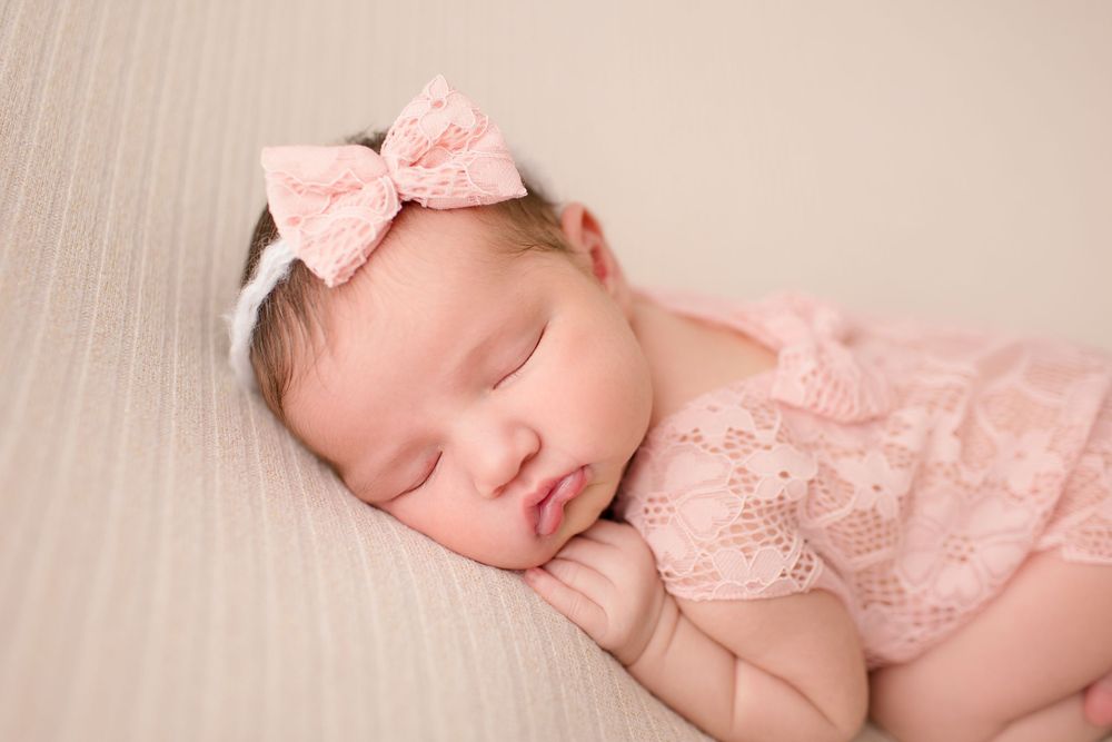 Peaceful sleeping newborn wearing pink lace romper and bow on cream background.