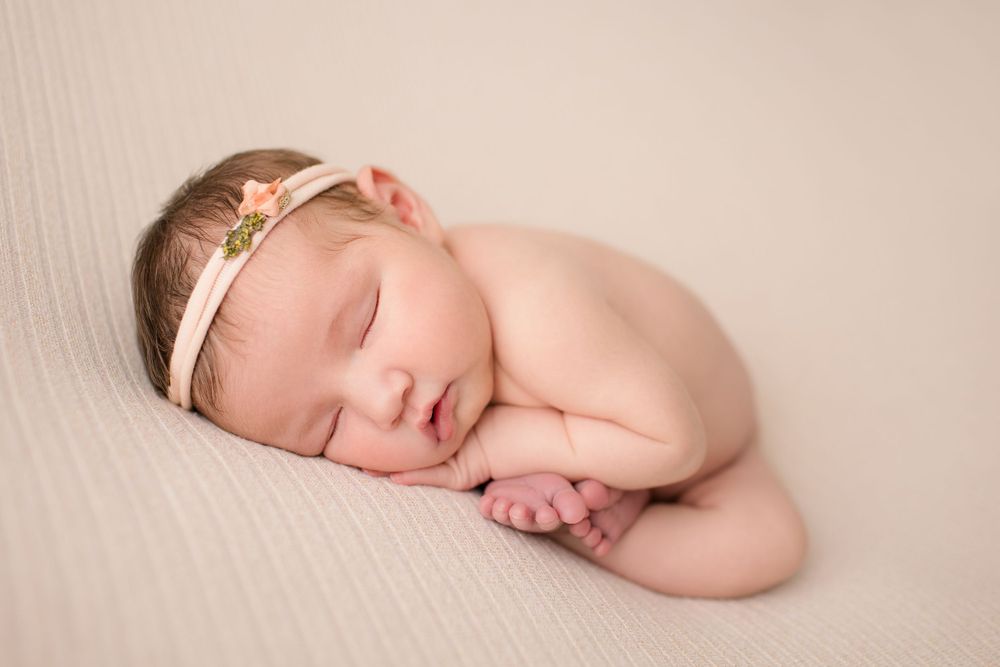 Peaceful newborn baby sleeping with gold headband on cream colored backdrop.
