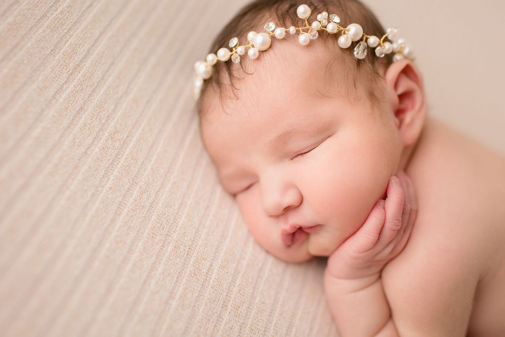 Sleeping newborn with floral crown resting hand under cheek on beige backdrop.