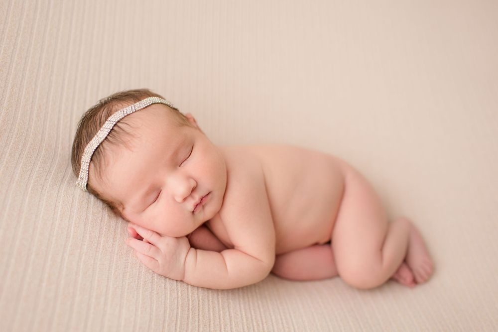 Sleeping newborn wearing white headband posed on textured beige background.