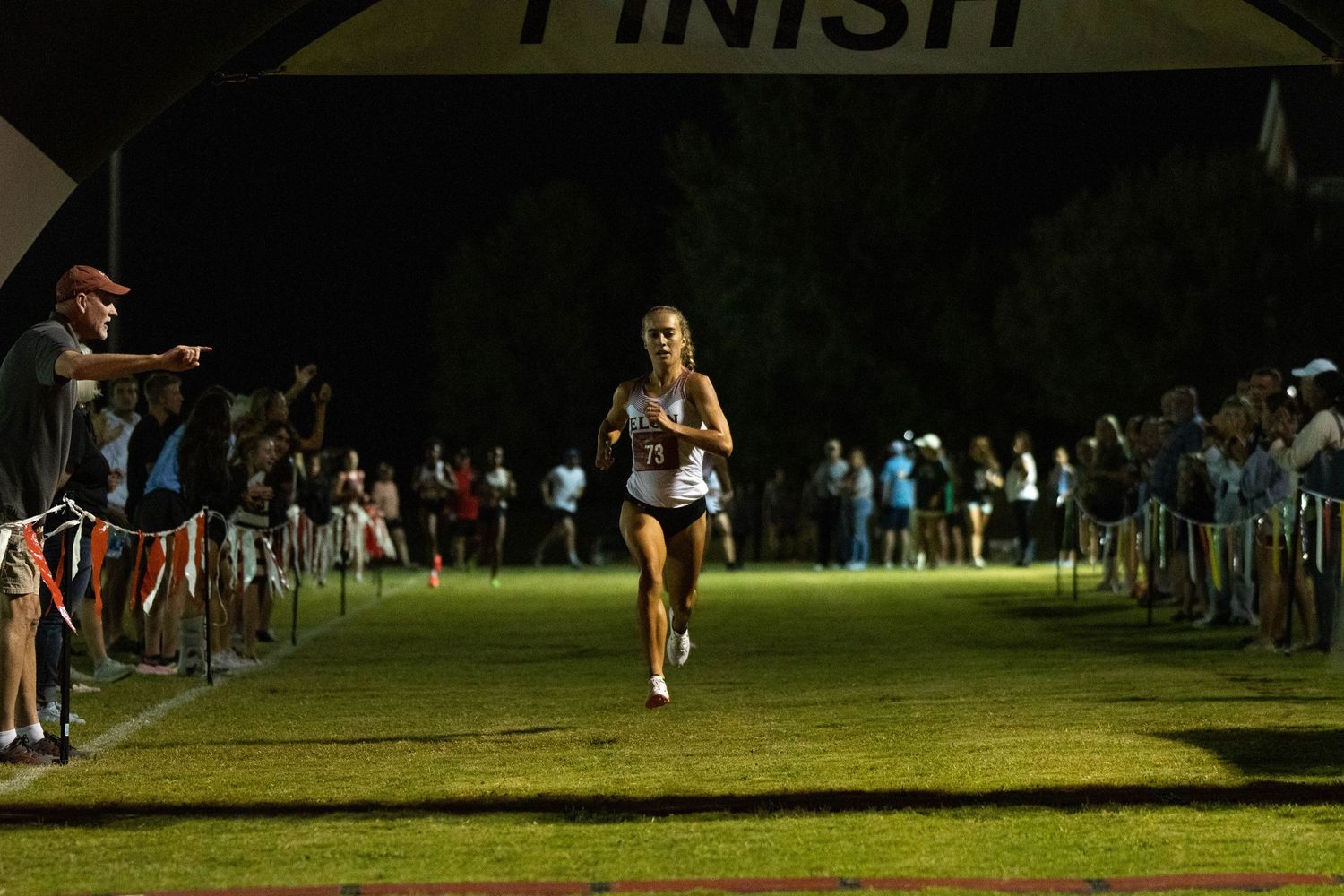 A nighttime cross country race with a runner approaching the finish line as spectators cheer.
