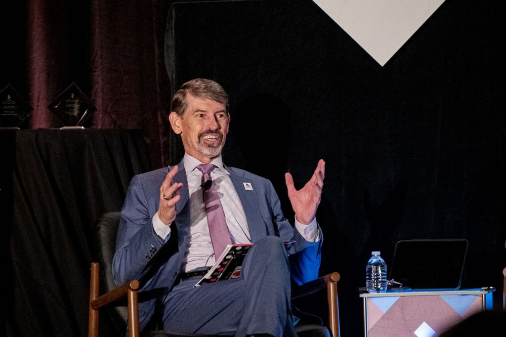 A speaker gestures while giving a presentation on stage in a dark auditorium.