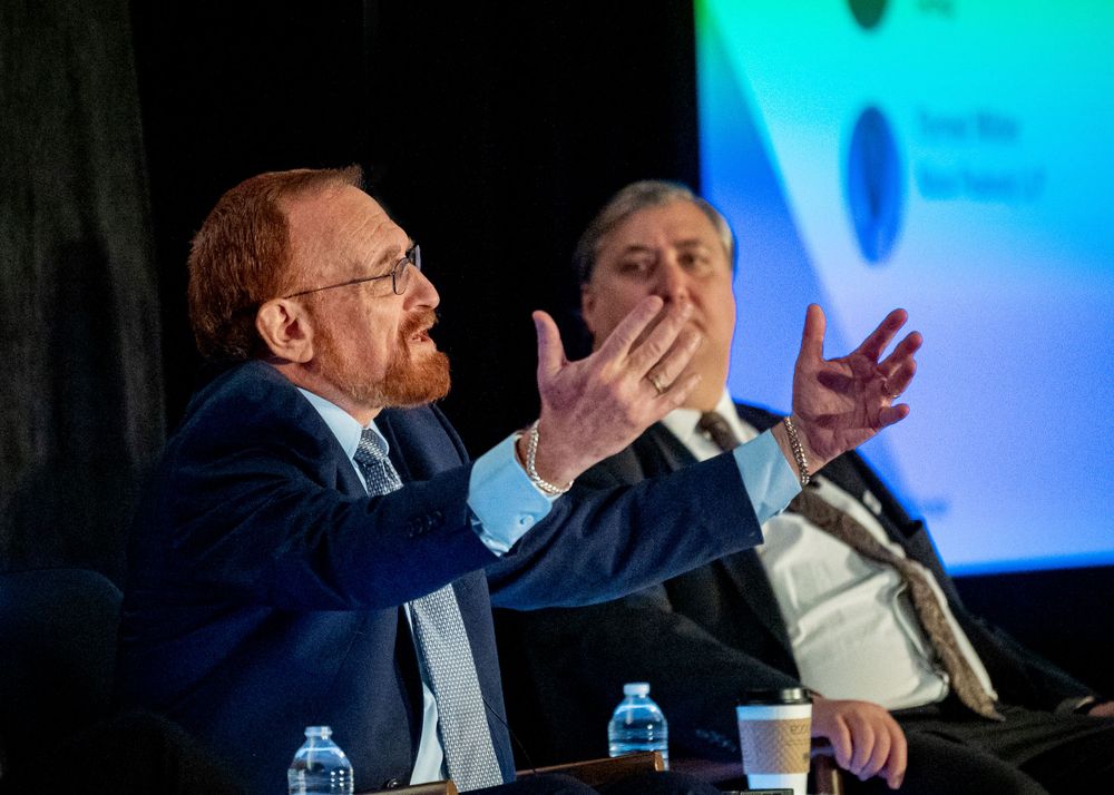 A speaker gestures animatedly while participating in a panel discussion with colorful projections behind them.
