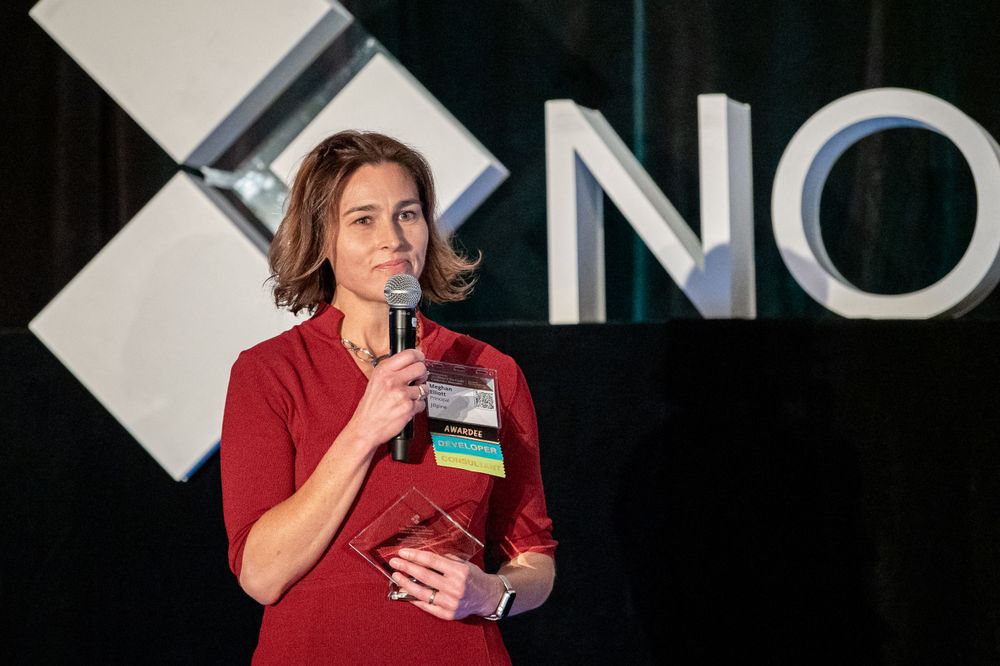 A speaker in red presents at a corporate event with the Novogradac logo displayed behind them.