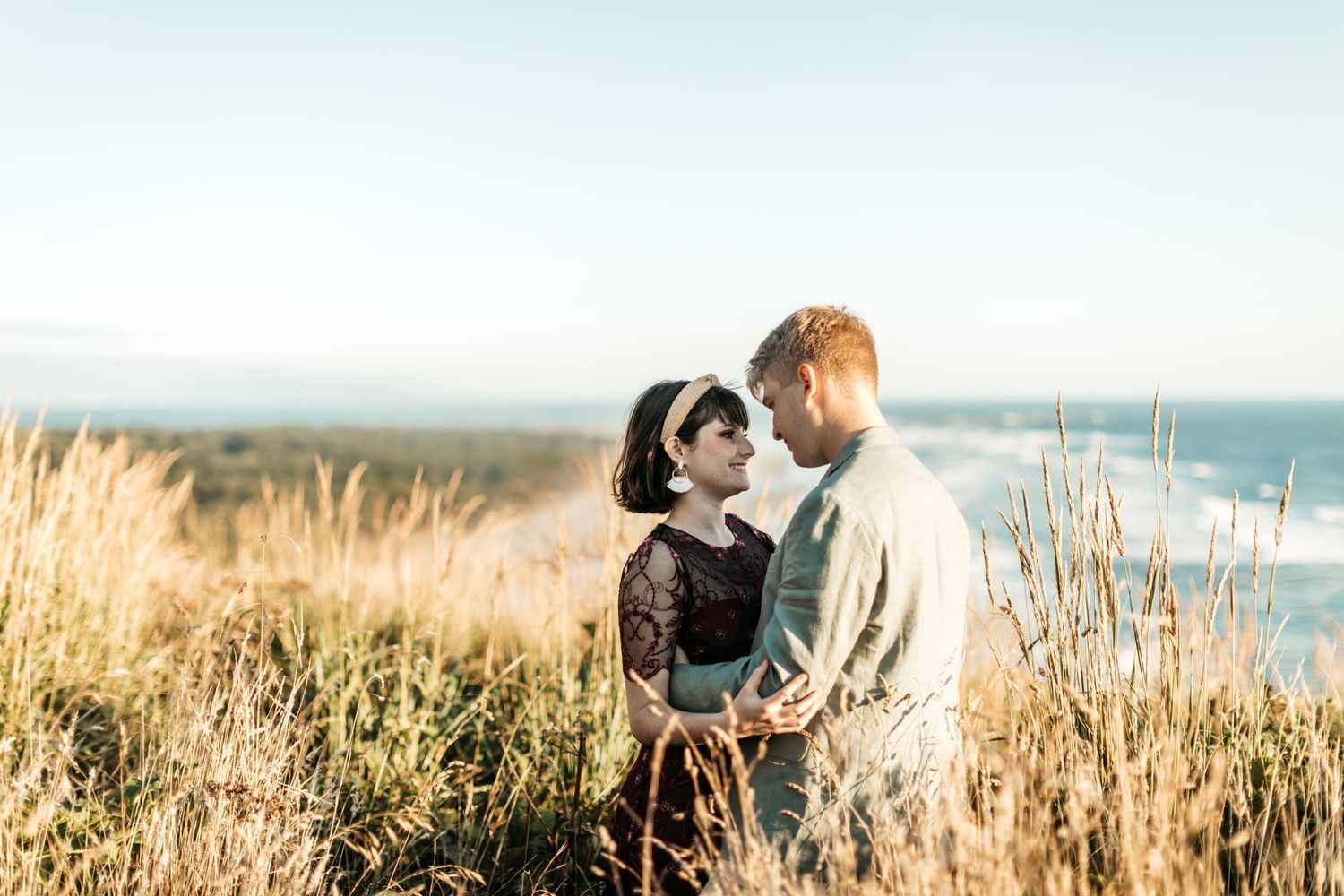 Girl and boy looking at each other while smiling and standing in beach grass on top of a hill with ocean in the distance
