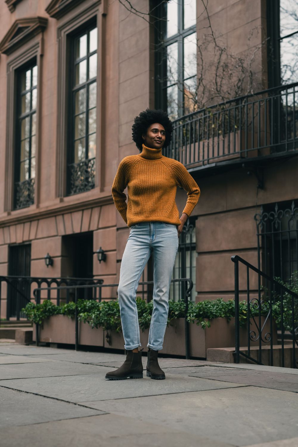 Figure in mustard sweater and light jeans standing on brownstone steps.