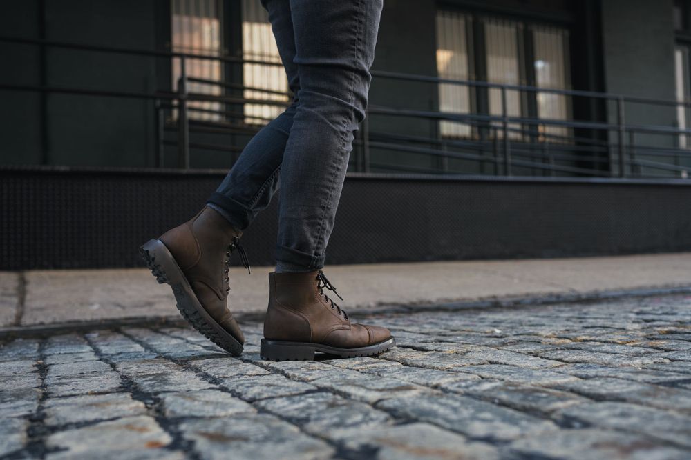 Feet walking on cobblestone street wearing dark jeans and brown boots.
