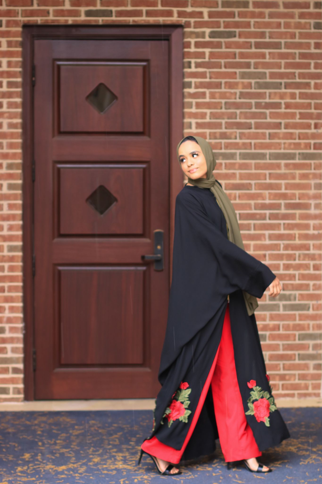 Elegant black abaya with red floral embroidery against a brick wall with wooden door.