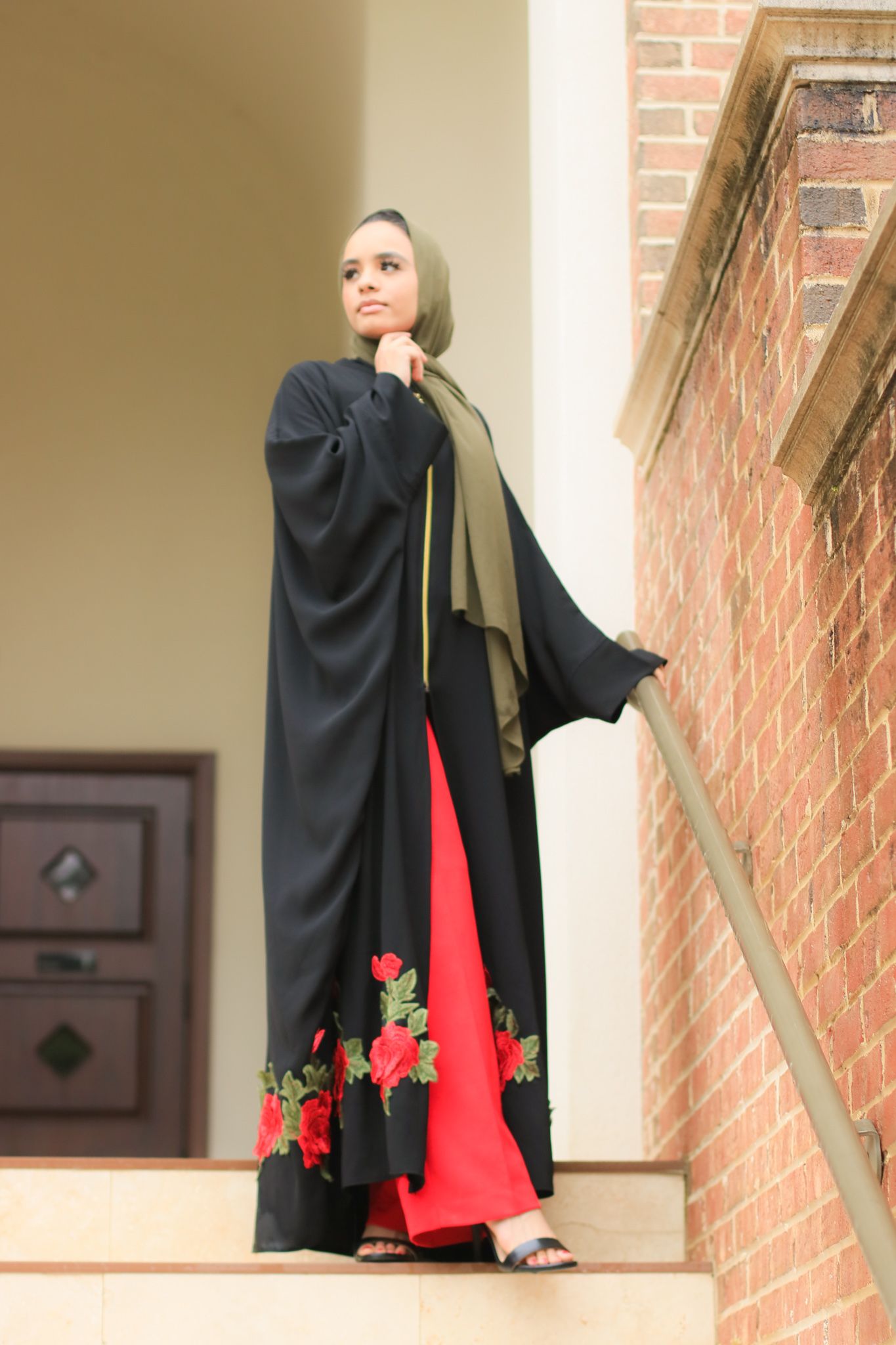 Flowing black abaya with red floral details shown on stone steps against brick wall.